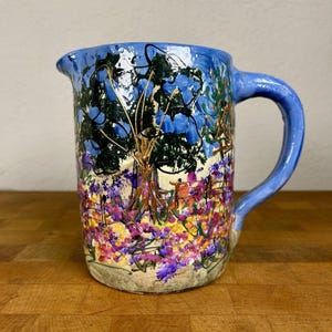 May include: A ceramic pitcher featuring a hand-painted landscape scene with a large tree, blue sky, and a vibrant field of purple, yellow, and pink flowers. The handle and rim are painted in a solid light blue, and the pitcher has a spout.