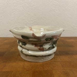 May include: A ceramic bowl with a wide, flared rim featuring two small pouring spouts. The exterior and interior have a glossy off-white glaze with irregular, abstract splatters of dark green, brown, and muted red colors across the surface.