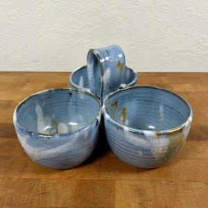 May include: A ceramic triple bowl server featuring three connected round compartments with a central arched handle. The bowls have a glossy light blue glaze with visible white and brown mottled accents and a textured ribbed exterior finish.