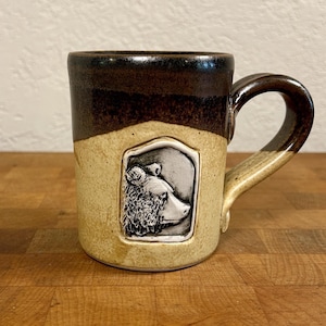 May include: A ceramic mug featuring a two-tone glaze with a dark brown upper section and a speckled cream lower section. A sculpted, rectangular relief of a bear head in profile is centered on the front, framed by a raised border.