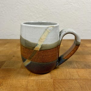 May include: A ceramic mug featuring a multi-toned horizontal color block design in shades of white, tan, brown, and dark gray. A diagonal, textured light-colored stripe cuts across the body, and the handle matches the earthy color palette.