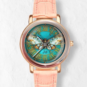 May include: A round wristwatch with a teal face featuring a butterfly design and Roman numerals. The watch has a gold-colored case and a light pink leather band. The watch face also has the initials "J.K."