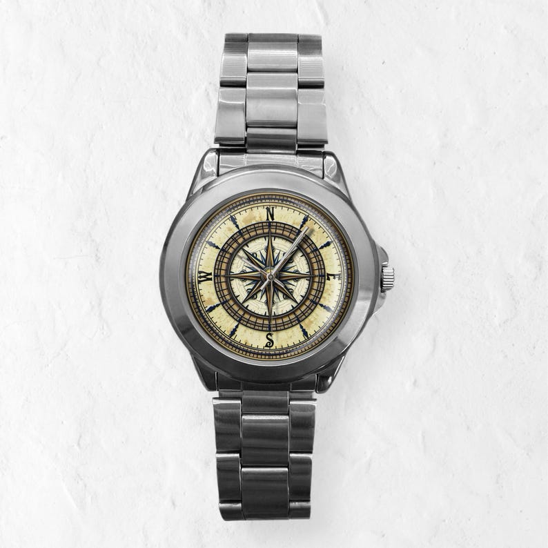 May include: A stainless steel wristwatch with a compass design on the face. The watch features a silver band and a cream-colored face with a detailed compass rose in shades of brown and gold. The watch is a classic nautical accessory.