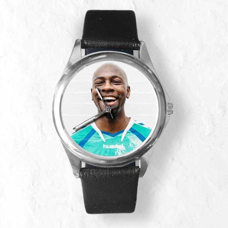 May include: A silver watch with a black leather band. The watch face features a photo of a smiling man wearing a teal and blue sports jersey with the word "hummel" printed on it.