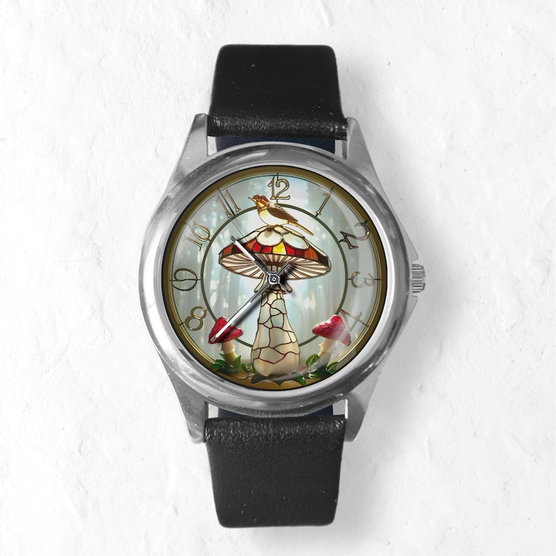 Puede incluir: Un reloj de pulsera con caja plateada y correa de cuero negro. La esfera presenta un dise&ntilde;o caprichoso de una seta con un p&aacute;jaro encima, sobre un fondo de bosque. El reloj tiene una pantalla anal&oacute;gica cl&aacute;sica.