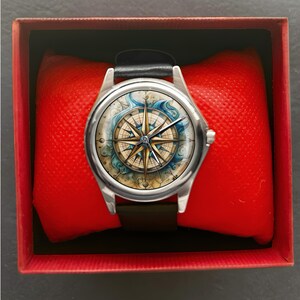 May include: A silver wristwatch with a black leather band, featuring a compass rose design on the watch face. The watch is presented in a red box with a red cushion.