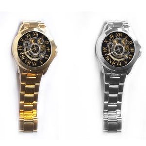 K&ouml;nnte beinhalten: Zwei Armbanduhren mit schwarzen Zifferbl&auml;ttern und goldenen r&ouml;mischen Ziffern als Stundenmarkierungen. Die Uhr links hat ein goldenes Armband, die Uhr rechts ein silbernes Armband.