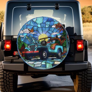 May include: A spare tire cover featuring a stained-glass design of a blue Jeep with a red roll bar, set against a mountain landscape with a yellow sun and a flying bird. The cover is mounted on a black Jeep.