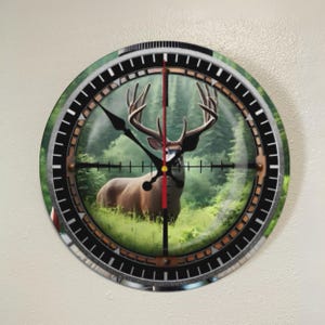 May include: Round wall clock featuring a deer in a forest scene, viewed through a scope-like design. The clock face has a black and silver bezel, with a red vertical line and crosshairs. The clock hands are black.