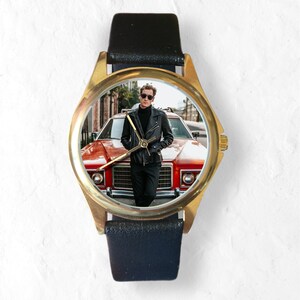 May include: A gold-tone wristwatch with a black leather band. The watch face features a photo of a person wearing a black leather jacket standing in front of a red car.