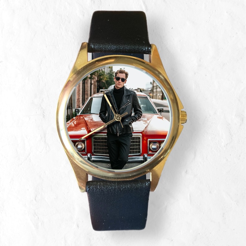 May include: A gold-tone wristwatch with a black leather band. The watch face features a photo of a person wearing a black leather jacket standing in front of a red car.