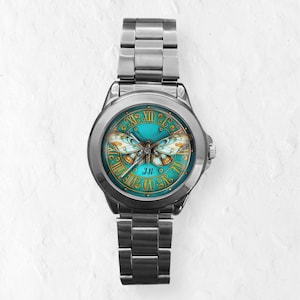 May include: A silver-toned wristwatch with a teal face featuring a butterfly design. The watch face has gold Roman numerals and a butterfly illustration with orange and white wings. The watch band is made of silver-toned metal.