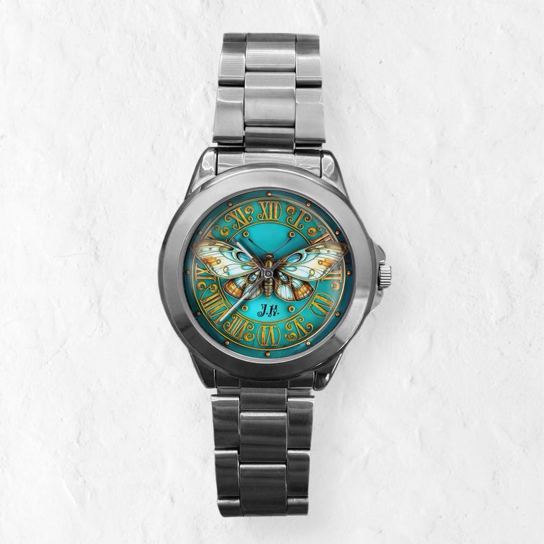 May include: A silver-toned wristwatch with a teal face featuring a butterfly design. The watch face has gold Roman numerals and a butterfly illustration with orange and white wings. The watch band is made of silver-toned metal.