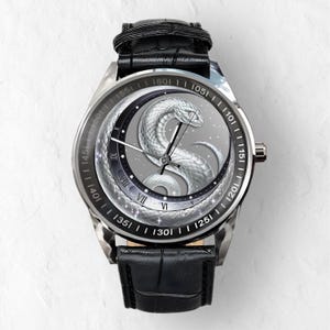 Puede incluir: Un reloj plateado con una correa de cuero negro. La esfera del reloj presenta un dise&ntilde;o de serpiente plateada con una luna y estrellas en el fondo. Los n&uacute;meros 101, 105, 110, 115, 120, 125, 130, 135, 140, 145, 150, 155, 160 est&aacute;n impresos alrededor del borde de la esfera del reloj.