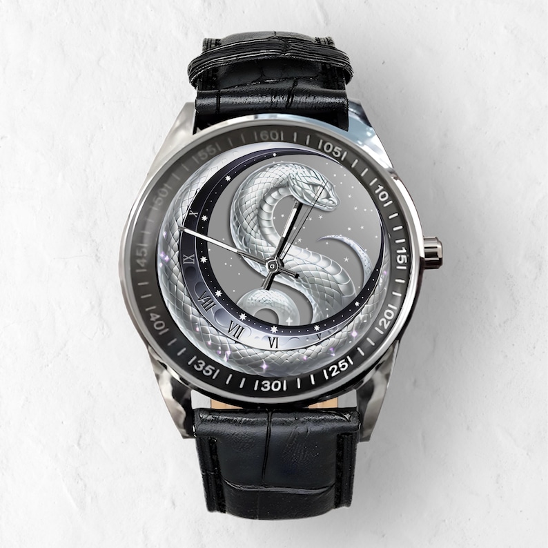 Puede incluir: Un reloj plateado con una correa de cuero negro. La esfera del reloj presenta un dise&ntilde;o de serpiente plateada con una luna y estrellas en el fondo. Los n&uacute;meros 101, 105, 110, 115, 120, 125, 130, 135, 140, 145, 150, 155, 160 est&aacute;n impresos alrededor del borde de la esfera del reloj.