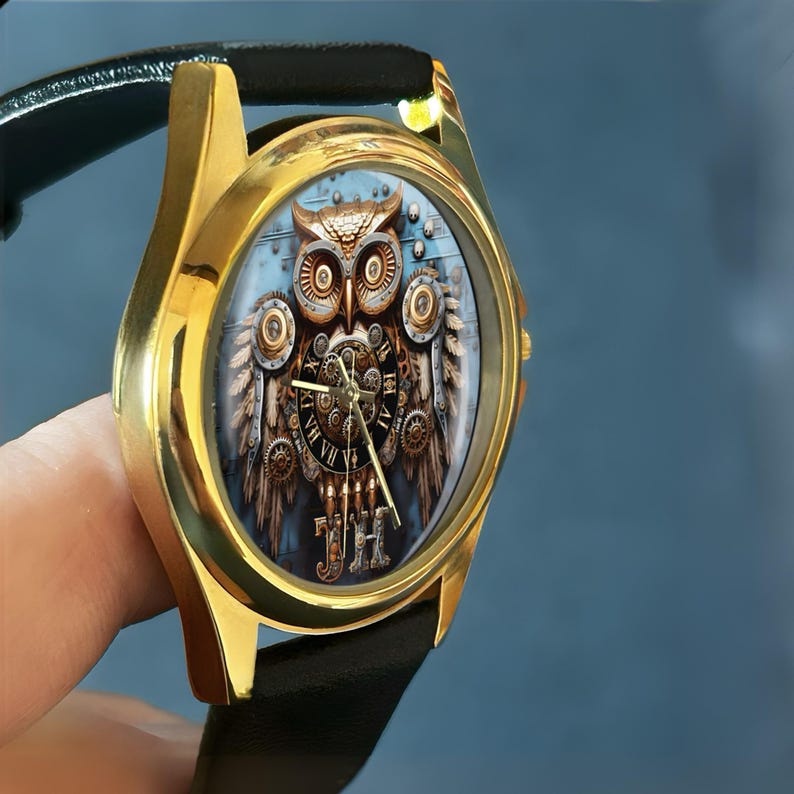 K&ouml;nnte beinhalten: Goldfarbene Armbanduhr mit schwarzem Lederarmband. Das Zifferblatt zeigt ein detailliertes Steampunk-Eulen-Design mit Zahnr&auml;dern und den Initialen "JH". Der Hintergrund ist hellblau.