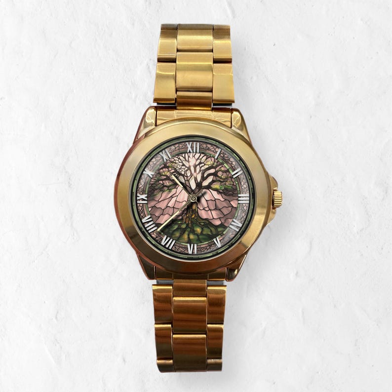 May include: Gold-toned wristwatch with a detailed tree of life design on the face. The watch features Roman numeral hour markers and a matching gold band. The watch face showcases a tree with pink and green hues.