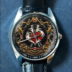 May include: A wristwatch with a black leather strap and a silver-tone metal case. The watch face features a complex mechanical gear design in gold and black, centered around a stylized white heart shape filled with small red gears and red accents.
