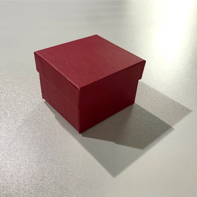K&ouml;nnte beinhalten: Eine kleine, quadratische, rote Geschenkbox mit strukturierter Oberfl&auml;che. Die Box ist geschlossen und wirft einen Schatten auf eine wei&szlig;e Oberfl&auml;che. Geeignet f&uuml;r Schmuck oder kleine Geschenke.