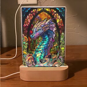 K&ouml;nnte beinhalten: Dekorative LED-Lampe mit einem farbenfrohen Drachendesign im Stil eines Glasfensters. Der Drache hat blaue, violette und gr&uuml;ne Schuppen, vor dem Hintergrund eines Waldes mit Pilzen. Die Lampe steht auf einem Holzsockel.
