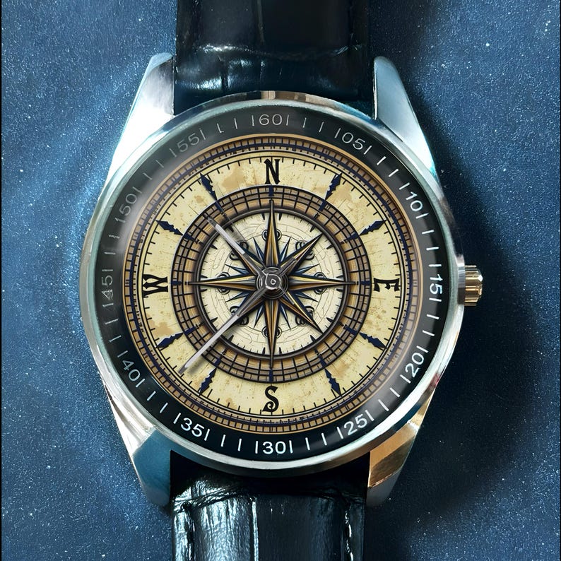 May include: A silver wristwatch with a black leather band. The watch face features a compass design with a beige background, black and gold accents, and the cardinal directions. The watch has a gold-colored crown.