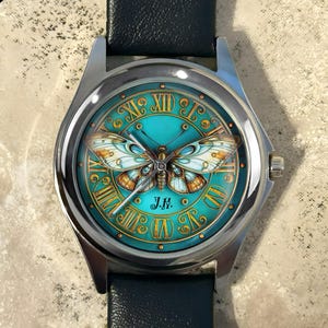 May include: A silver wristwatch with a teal face featuring a detailed butterfly design. The watch face has gold Roman numerals and a black leather band. The watch face also has the initials "J.K."