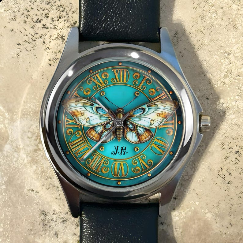 May include: A silver wristwatch with a teal face featuring a detailed butterfly design. The watch face has gold Roman numerals and a black leather band. The watch face also has the initials "J.K."