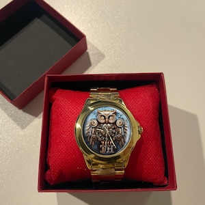 K&ouml;nnte beinhalten: Goldfarbene Armbanduhr mit detailliertem Eulen-Design auf dem Zifferblatt, pr&auml;sentiert in einer roten Box. Die Eulen-Illustration zeigt Zahnr&auml;der und mechanische Elemente vor blauem Hintergrund. Die Uhr hat ein goldenes Armband und liegt auf einem roten Kissen.