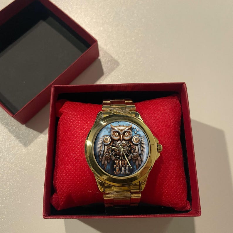 K&ouml;nnte beinhalten: Goldfarbene Armbanduhr mit detailliertem Eulen-Design auf dem Zifferblatt, pr&auml;sentiert in einer roten Box. Die Eulen-Illustration zeigt Zahnr&auml;der und mechanische Elemente vor blauem Hintergrund. Die Uhr hat ein goldenes Armband und liegt auf einem roten Kissen.