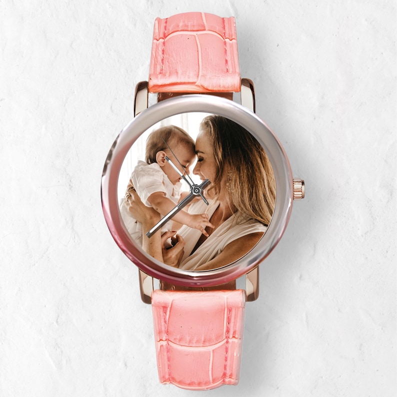 May include: A pink leather wristwatch with a silver face featuring a photo of a mother and child. The watch has a rose gold colored case and hands.