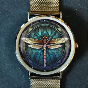 May include: A gold-toned wristwatch with a mesh band. The watch face features a colorful dragonfly design with shades of gold, orange, and teal. The watch has a white bezel and a gold-colored case.