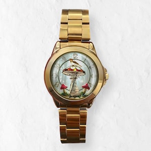 Puede incluir: Un reloj de pulsera dorado con una esfera detallada que presenta un dise&ntilde;o de hongo con un p&aacute;jaro posado encima. El reloj tiene una correa dorada y una esfera redonda. La seta tiene detalles en rojo y marr&oacute;n, y la esfera tiene un fondo de bosque.