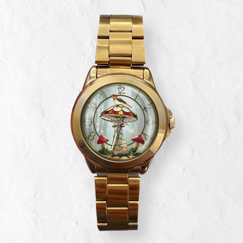 Puede incluir: Un reloj de pulsera dorado con una esfera detallada que presenta un dise&ntilde;o de hongo con un p&aacute;jaro posado encima. El reloj tiene una correa dorada y una esfera redonda. La seta tiene detalles en rojo y marr&oacute;n, y la esfera tiene un fondo de bosque.