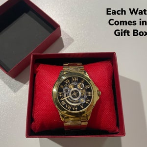 K&ouml;nnte beinhalten: Eine goldfarbene Uhr mit einem Skelettzifferblatt und r&ouml;mischen Ziffern als Stundenmarkierungen. Die Uhr befindet sich in einer roten Geschenkbox. Jede Uhr wird in einer Geschenkbox geliefert.