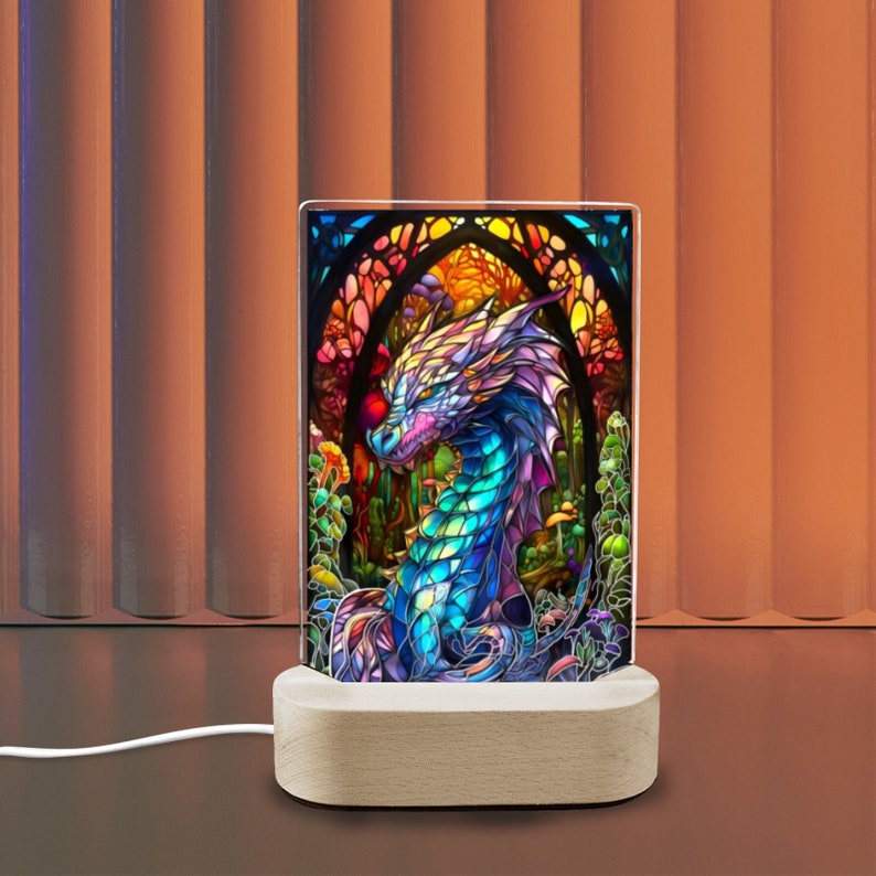 K&ouml;nnte beinhalten: Ein rechteckiges Leuchtpaneel zeigt eine farbenfrohe Drachenillustration. Der Drache ist in Blau-, Lila- und Gr&uuml;nt&ouml;nen dargestellt, vor einem Hintergrund im Buntglasstil. Das Paneel steht auf einem Holzsockel, mit einem wei&szlig;en Kabel.