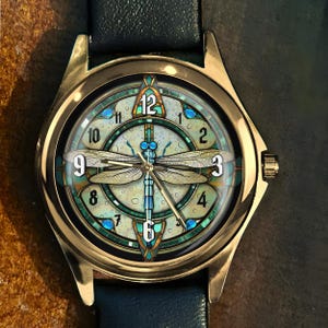 May include: A gold-toned wristwatch with a detailed dragonfly design on the face. The watch features a blue leather band and a round face with Roman numerals. The dragonfly is centered, with intricate wing details and blue accents.