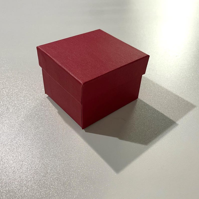 May include: A small, square, red gift box with a textured surface. The box has a lid and a base, and it casts a shadow on the white surface below. The box is likely used for jewelry or small gifts.
