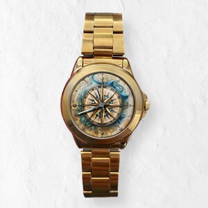 May include: Gold-toned wristwatch featuring a detailed compass rose design on the face. The watch has a gold band and a round face with a nautical theme, including blue and beige accents. The watch is a stylish accessory.