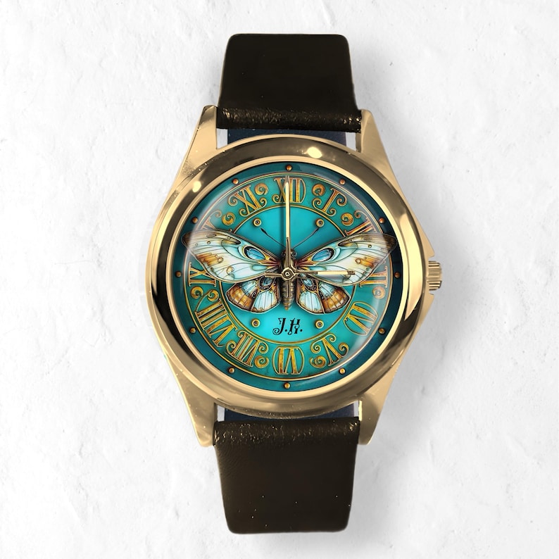 May include: Gold-tone wristwatch with a teal face featuring a butterfly design. The watch face has Roman numerals and the initials "J.H." The watch has a black leather band.