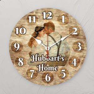 May include: A round wooden wall clock with a photograph of a couple, and the text "Hubbart's Home". The clock face has white numerals and hands. The background has a wood grain texture. The clock hands show the time.