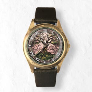 May include: A gold-toned wristwatch with a black leather band. The watch face features a detailed illustration of a tree with pink and green hues, set against a white background. Roman numerals mark the hours.