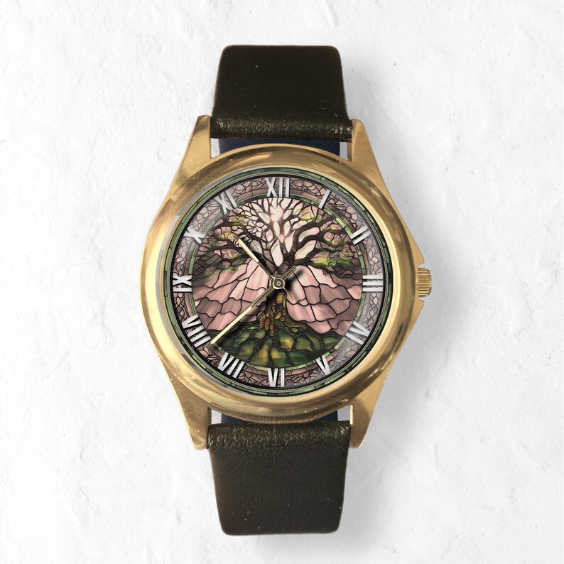 May include: A gold-toned wristwatch with a black leather band. The watch face features a detailed illustration of a tree with pink and green hues, set against a white background. Roman numerals mark the hours.