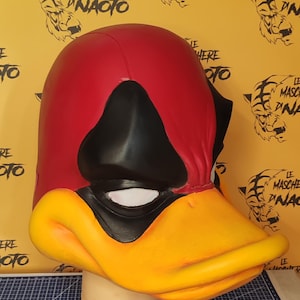 Può includere: Una maschera da anatra integrale con testa rossa, maschera per gli occhi nera e un grande becco giallo. La maschera è su una testa di manichino bianca. Lo sfondo è giallo con illustrazioni di tigri e testo.