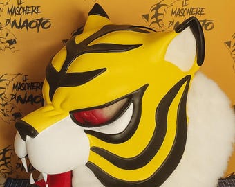 May include: A yellow and black tiger mask with red eyes and white teeth. The mask has a white fur trim and the text "PRODOTTO ESCLUSIVO" in a black circle. The background has the text "LE MASCHERE DINAOTO".