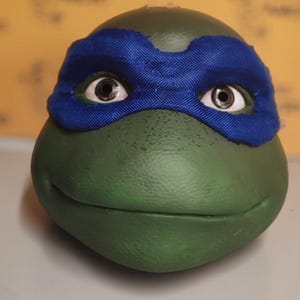 May include: A green, rounded character head resembling a turtle, featuring a textured surface and a smooth, closed mouth. The figure wears a bright blue fabric eye mask that covers the upper portion of the face, revealing two brown eyes.
