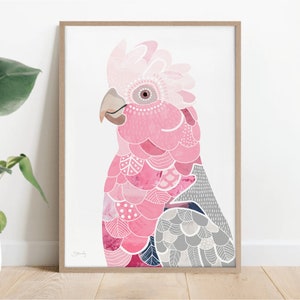 May include: A framed art print featuring a pink and gray parrot illustration. The bird has a detailed design with various shades of pink, gray, and white. The artwork is set against a white background and framed in a light brown wooden frame.