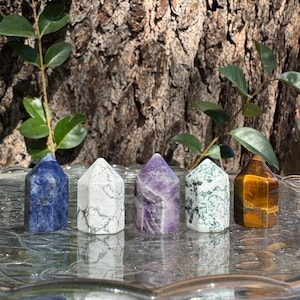 May include: Five gemstone crystal towers in a row, each with a different color and pattern. The crystals are blue, white with gray veining, purple, white with green inclusions, and brown with yellow stripes. The crystals are on a glass surface.