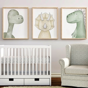 K&ouml;nnte beinhalten: Drei gerahmte Kunstdrucke mit Dinosauriern. Die Drucke zeigen Aquarellillustrationen eines Brontosaurus, Triceratops und T-Rex in sanften Gr&uuml;n- und Beiget&ouml;nen. Ein wei&szlig;es Babybett und ein grauer Sessel sind ebenfalls zu sehen.