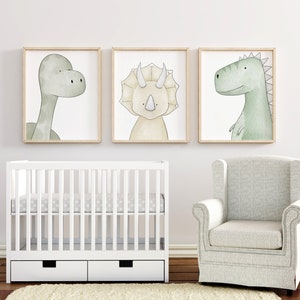 K&ouml;nnte beinhalten: Drei gerahmte Kunstdrucke mit Dinosauriern in sanften Gr&uuml;n- und Beiget&ouml;nen h&auml;ngen &uuml;ber einem wei&szlig;en Kinderbett. Ein passender Sessel befindet sich in der N&auml;he. Die Kunstwerke zeigen einen Brontosaurus, einen Triceratops und einen Tyrannosaurus Rex.