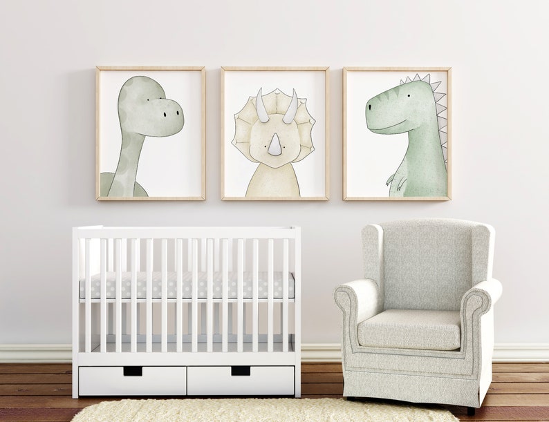 K&ouml;nnte beinhalten: Drei gerahmte Kunstdrucke mit Dinosauriern in sanften Gr&uuml;n- und Beiget&ouml;nen h&auml;ngen &uuml;ber einem wei&szlig;en Kinderbett. Ein passender Sessel befindet sich in der N&auml;he. Die Kunstwerke zeigen einen Brontosaurus, einen Triceratops und einen Tyrannosaurus Rex.